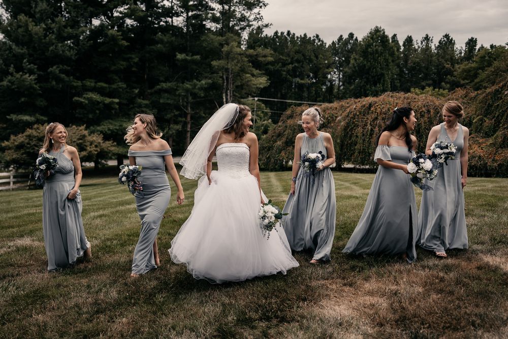 A bridal party in gray dresses and a white wedding gown pose together on a grassy hill with evergreen trees in the background.
