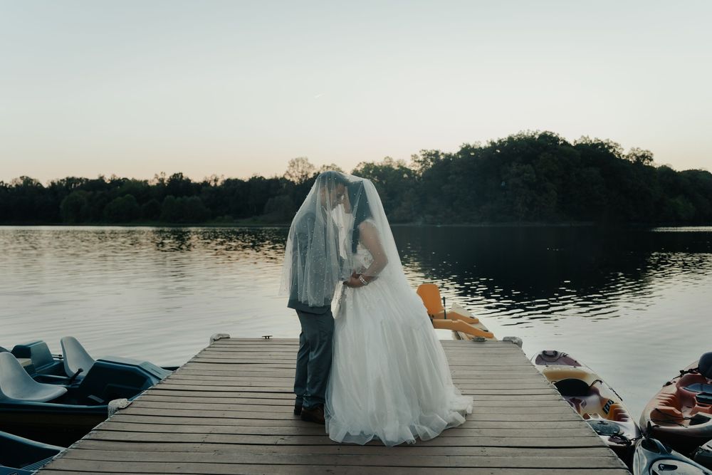 A romantic wedding couple stands on a wooden dock overlooking a serene lake at sunset.