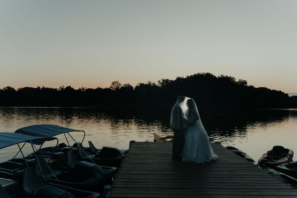 A romantic wedding couple stands on a wooden dock overlooking a serene lake at sunset.