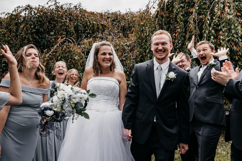 Wedding party in gray and black attire laughs together outdoors with willow trees in background.