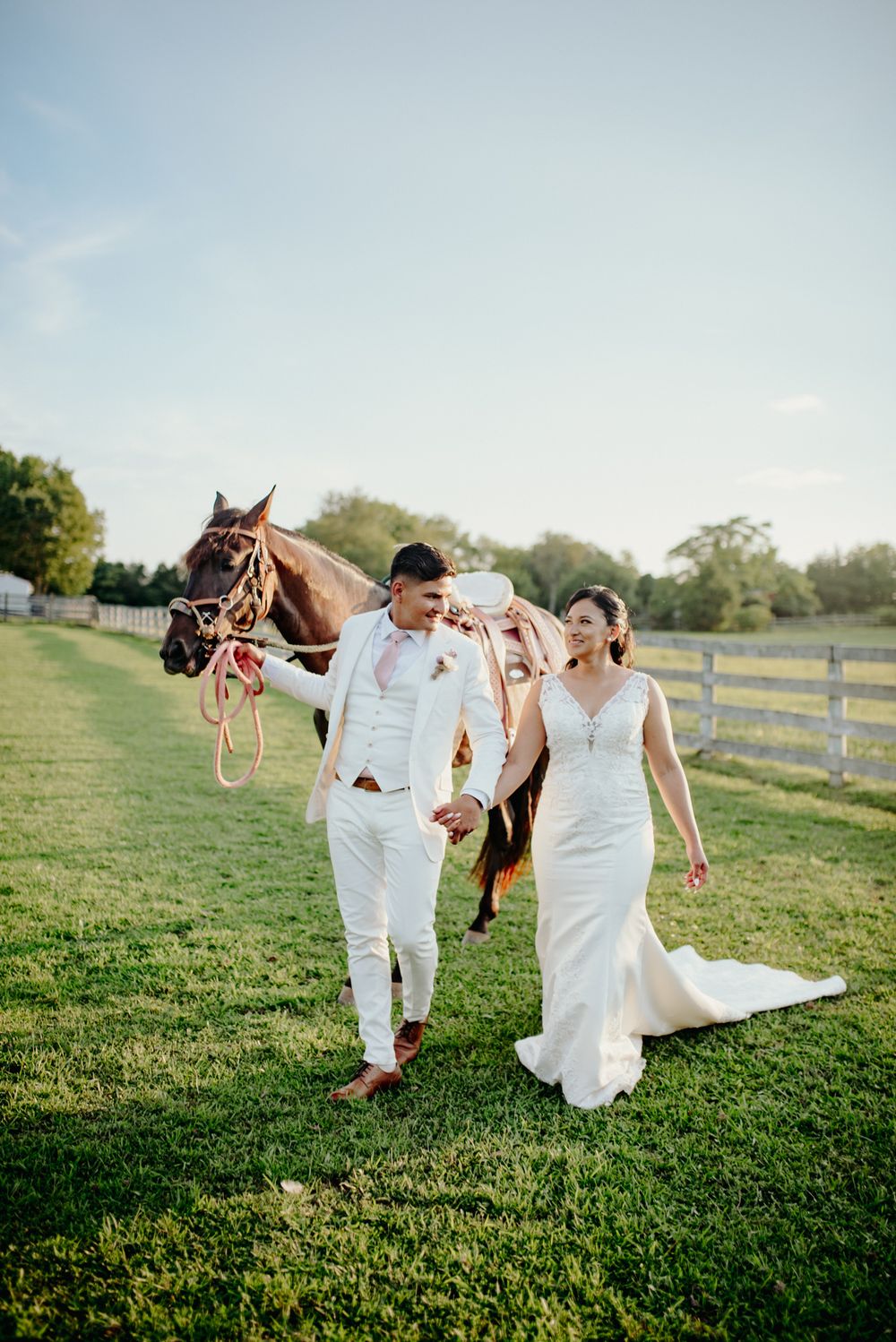 A couple in white wedding attire walk together on a grassy field with a horse and wooden fence at sunset.