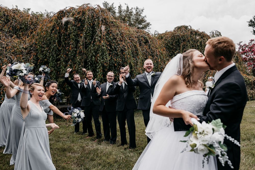 Wedding party in formal attire celebrates outdoors as newlyweds share romantic moment with bridal bouquet.
